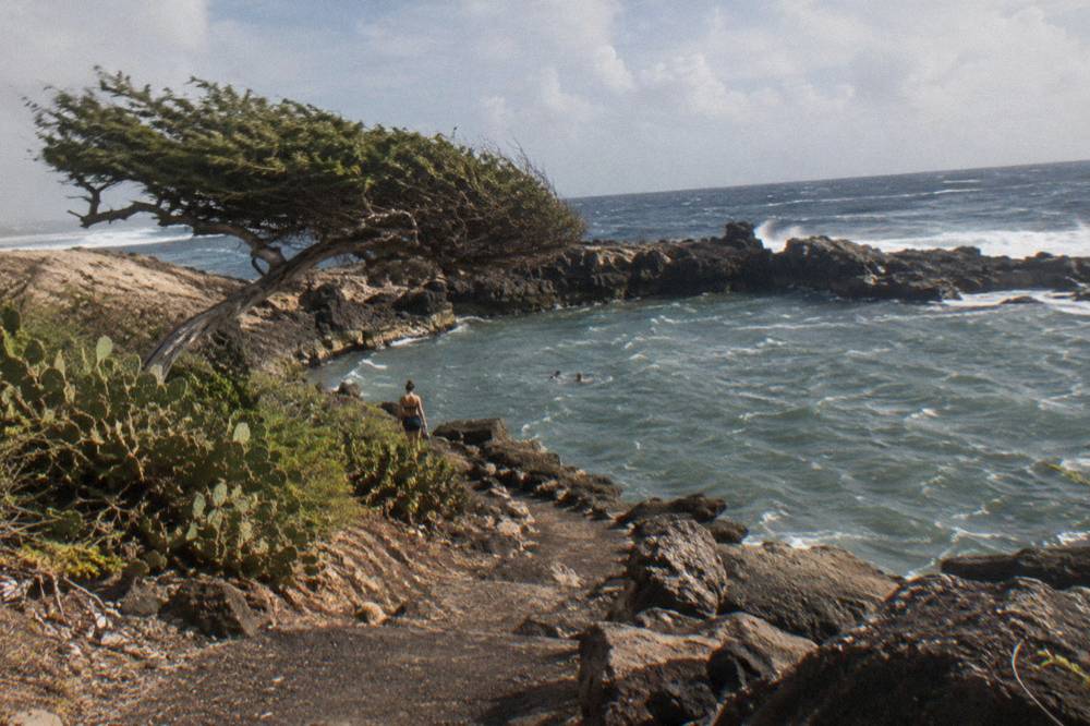 A rocky volcanic track descending toward a rock-ringed pool on a rugged coastline