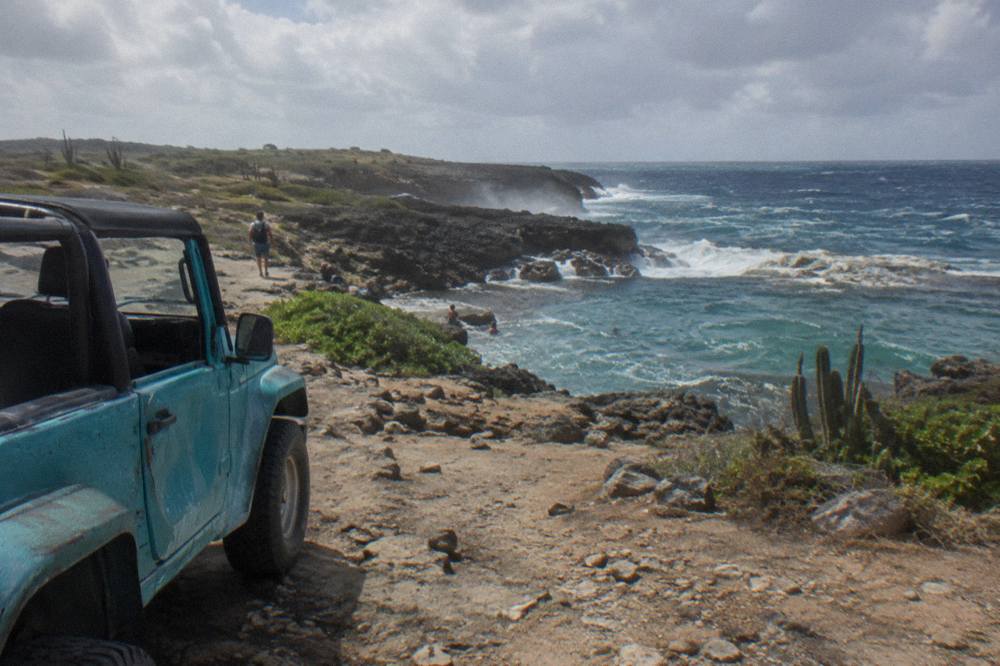 A jeep parked on a rocky dirt track above a rocky coastline with waves breaking against dark volcanic stone
