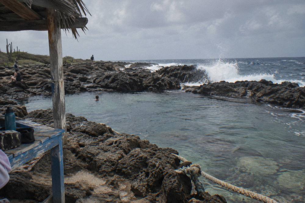 Waves breaking against dark volcanic rock at the edge of a natural pool with clear water inside the basin