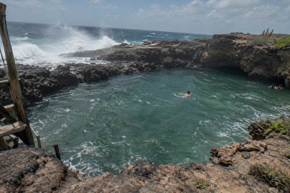 Natural pool enclosed by dark volcanic rock with waves breaking on the outer reef beyond