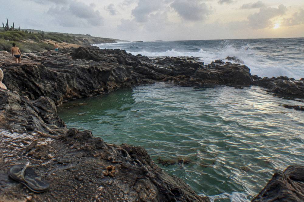 A natural rock pool set among dark volcanic boulders with ocean waves breaking beyond the rim