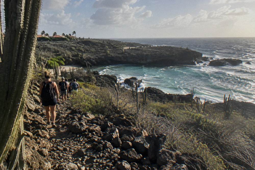 A guided group walking a rocky coastal trail through dry terrain toward a sheltered cove