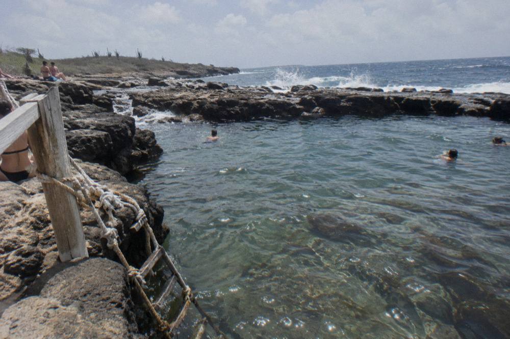 Clear shallow water in a rock-ringed pool with dark volcanic stone and gentle waves beyond the rim