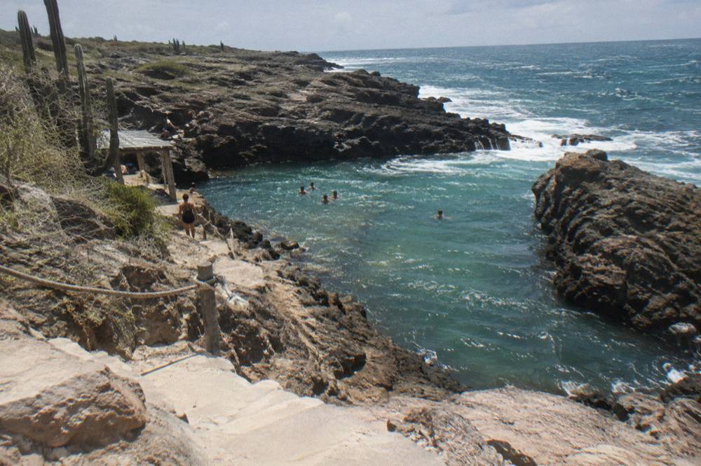Rocky volcanic terrain sloping toward a sheltered pool with pale sand-colored stone steps in the foreground