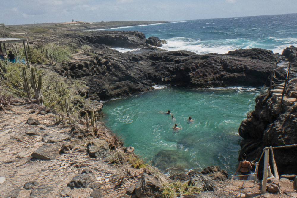 Dark volcanic rocks forming a natural basin with clear water, seen from a rocky trail above the coast
