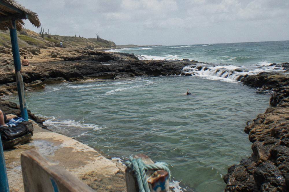 A natural rock pool enclosed by dark volcanic stone with gentle waves washing over the rim into calm water