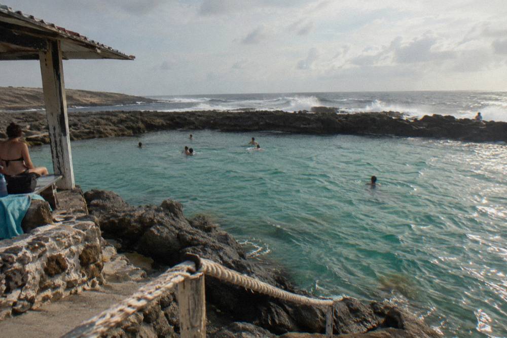 A calm turquoise rock pool ringed by dark volcanic stone with waves breaking beyond the rim