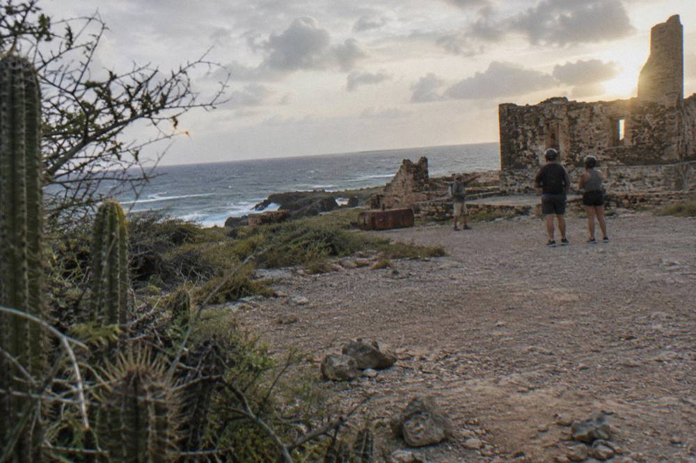 A rocky coastal gravel area with cactus, ocean surf, stone ruins and three people near the ruins at dusk