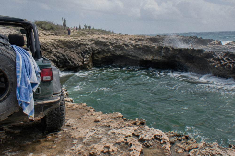 An open-air four by four vehicle standing on a rocky track above a volcanic rock pool on the coast