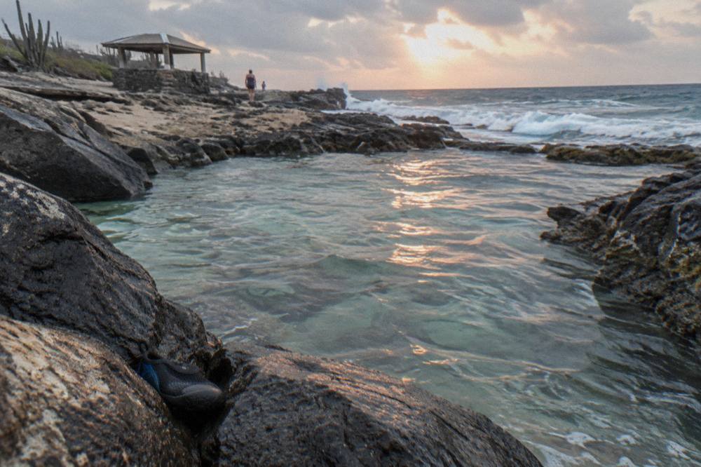 A natural rock pool enclosed by dark volcanic stone with clear water and ocean waves breaking on the outer rim