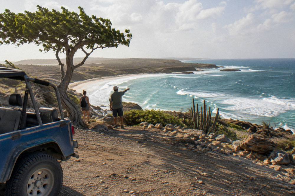 Open-air jeep parked on rocky ground near coastal cliffs with a guide pointing toward the shoreline