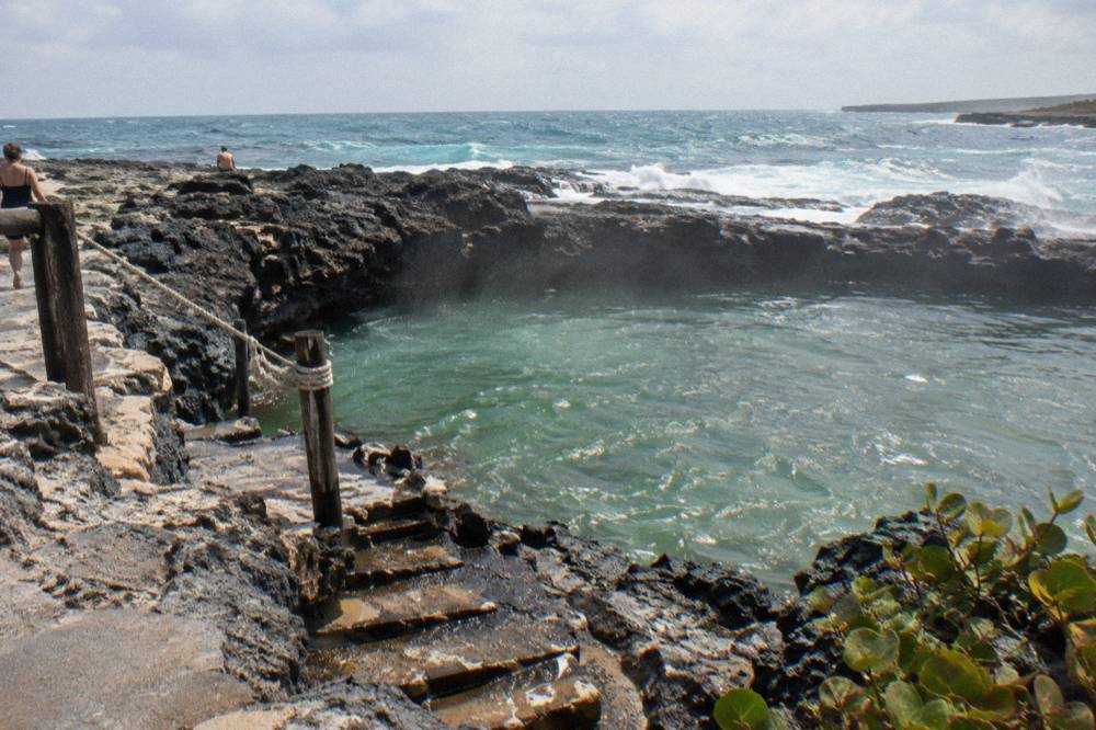 A natural rock pool enclosed by dark volcanic stone with ocean waves breaking beyond