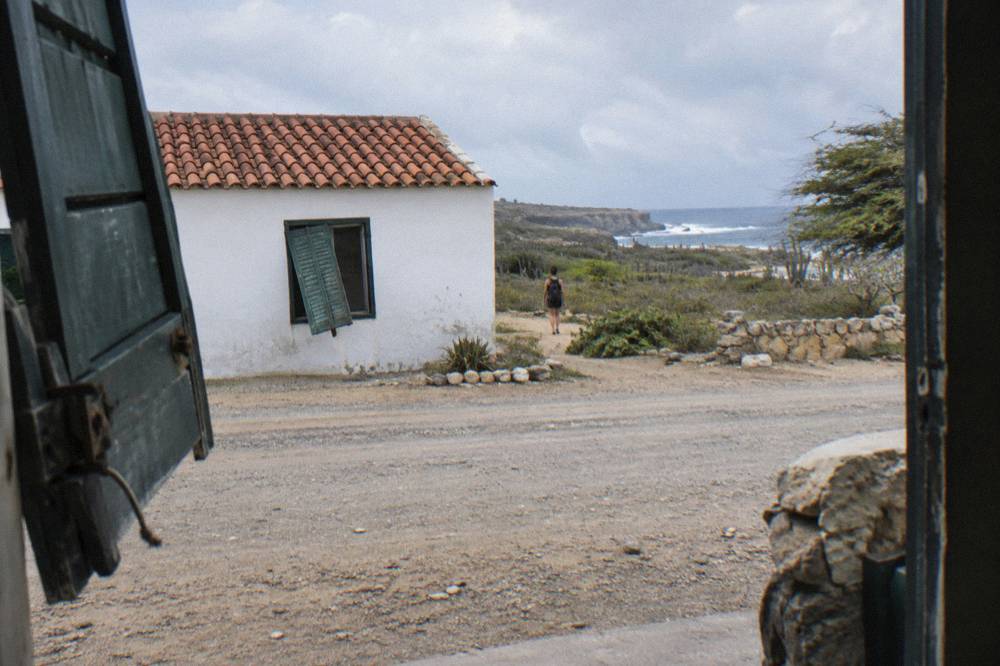 A white building with a red tile roof and crooked green shutter beside a dry gravel road, seen through a doorway