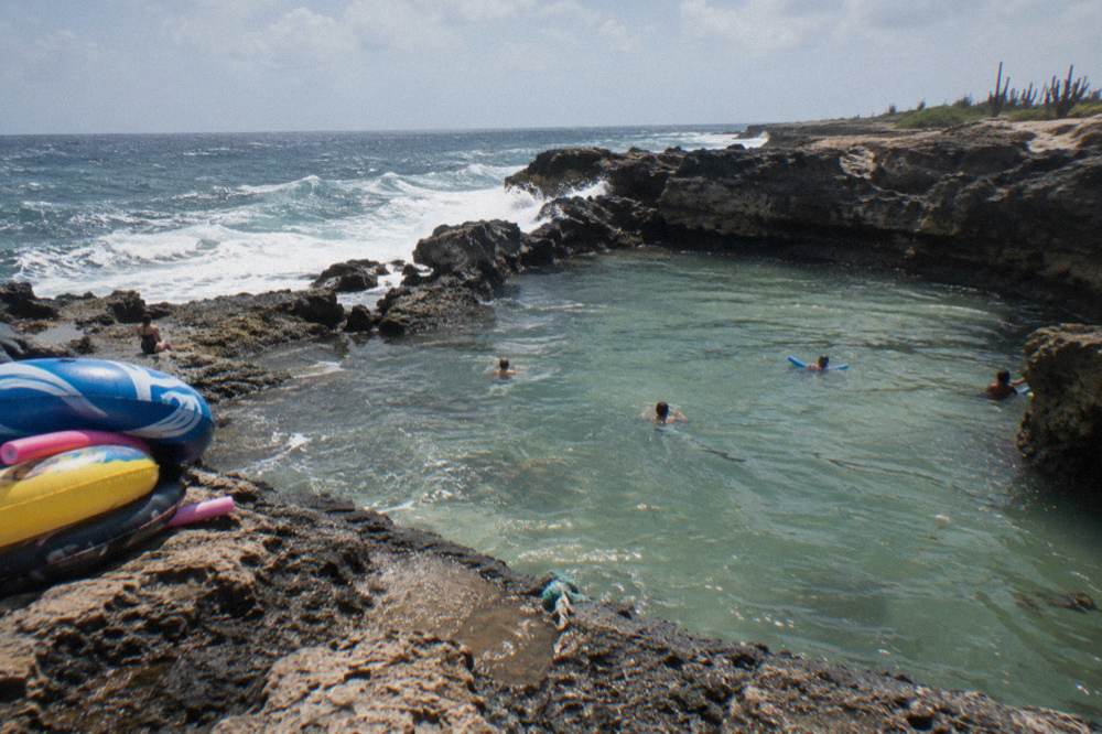 A natural rock pool enclosed by dark volcanic stone with waves breaking beyond it on the coast