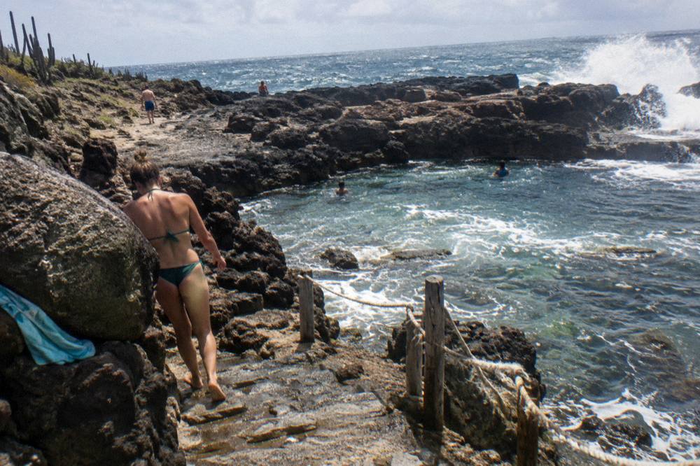 Visitor in swimwear walking on dark volcanic rock toward a coastal inlet