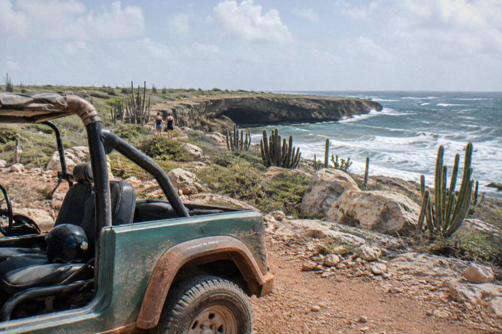 Open-air jeep parked on a rocky cactus-lined coastal track above cliffs and rough ocean surf