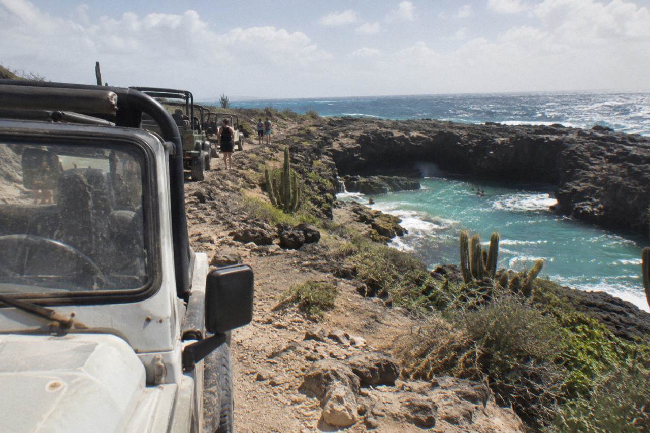 Tour jeeps parked on a rocky desert trail above a turquoise rock pool on Aruba's north-east coast