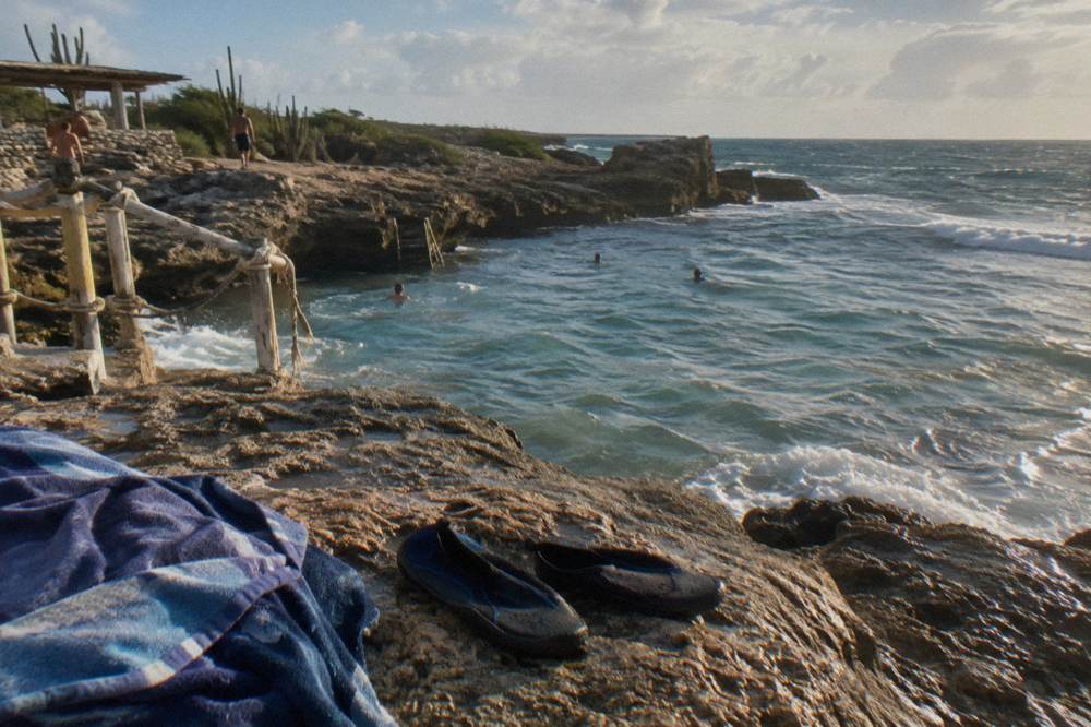 Towel And Water Shoes On A Rocky Ledge Above A Natural Pool With Swimmers And Ocean Surf