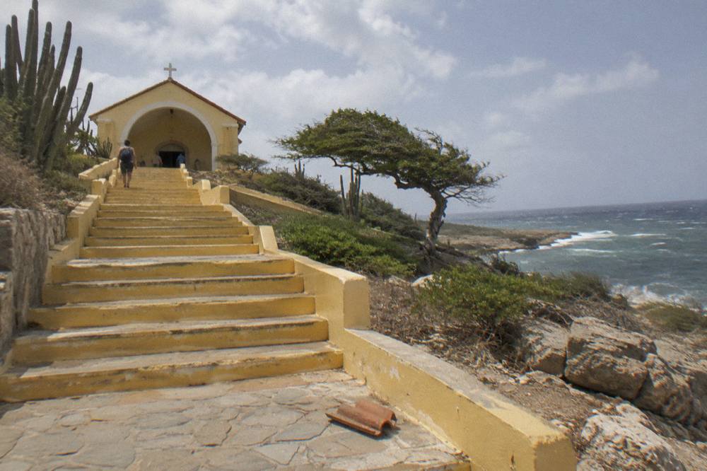 Yellow stairway up to a small chapel on an arid coastal hillside with cacti and ocean surf
