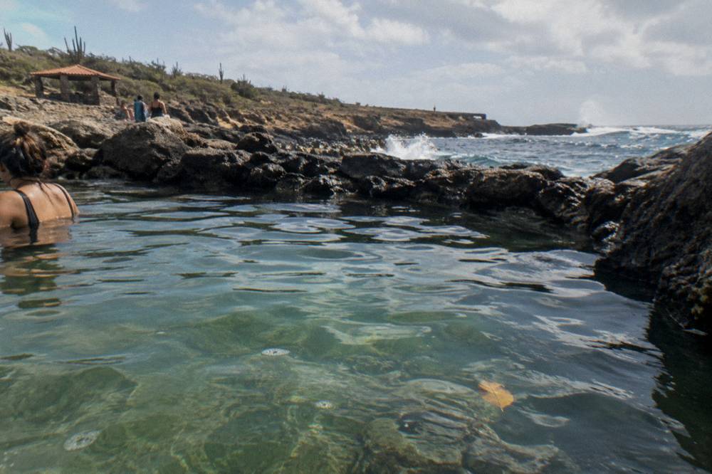 A calm natural rock pool enclosed by dark volcanic stone beside a crashing surf line