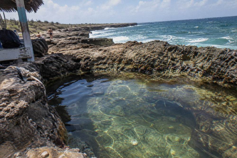 Clear shallow rock pool in rough coastal limestone beside ocean surf, with a small structure and people nearby