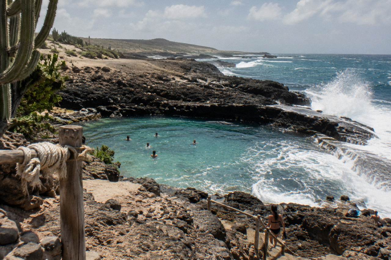 A natural rock pool set among dark volcanic stones on a coastline with waves breaking beyond the rim