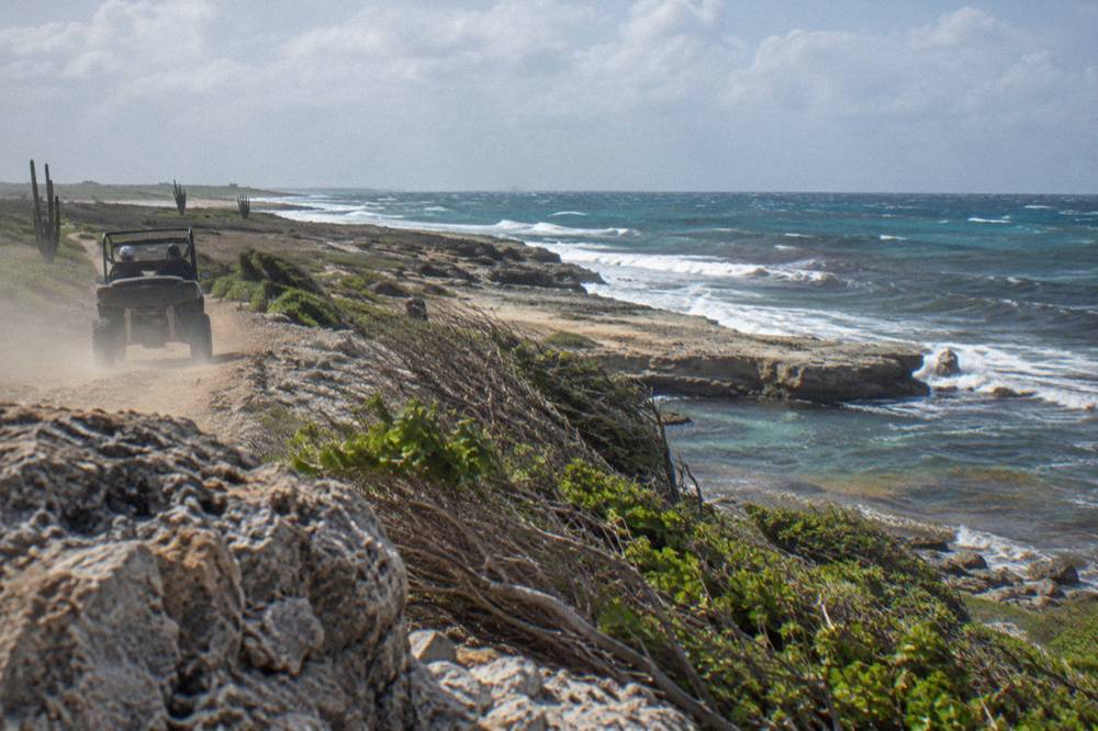 A single UTV buggy kicking up dust on a rocky coastal track with flat limestone terraces and open sea beyond