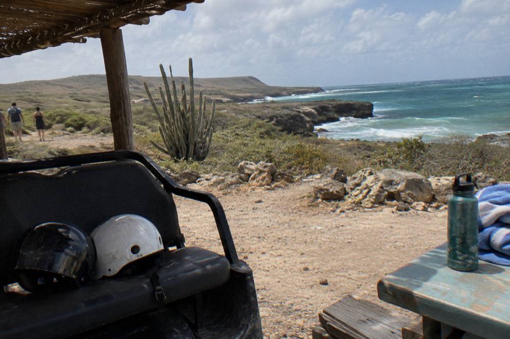 A shaded rest stop on a rocky trail with helmets resting on a UTV seat and calm turquoise water visible past the rocks