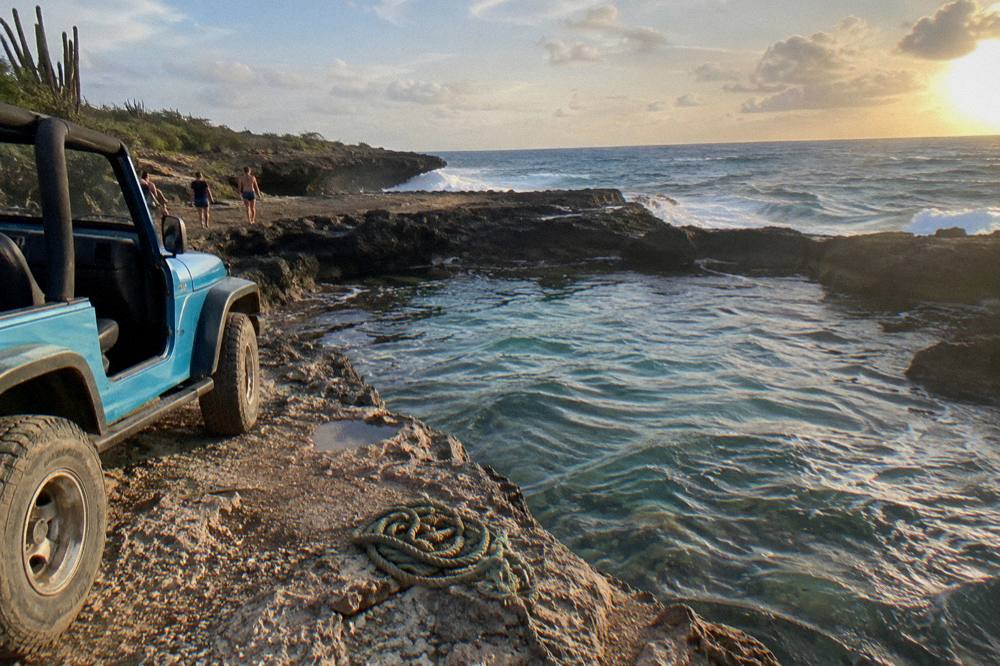 An open-air jeep parked beside a natural rock pool at the end of a rugged track, with clear shallow water and dark volcanic rock ledges