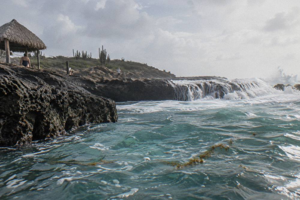 Turquoise water pooling inside a ring of dark volcanic rock on a rocky coastline, with white surf breaking on the outer rocks