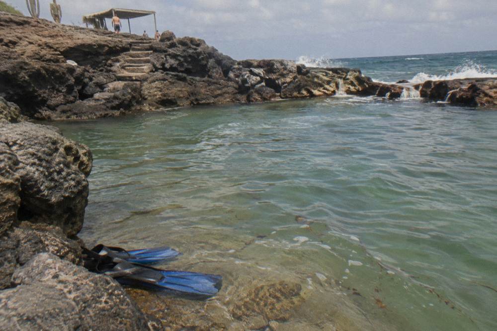 Calm turquoise water filling a rocky coastal basin, with a snorkeler's fins visible at the edge of dark stone