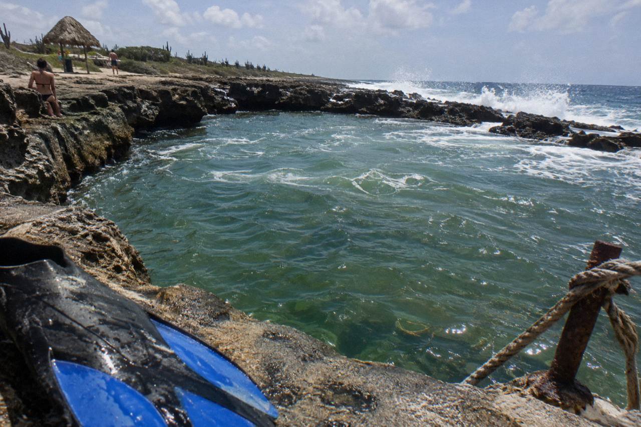 A natural rock pool set among dark volcanic cliffs on Aruba's rugged northeast coast, with waves breaking on the rocks beyond