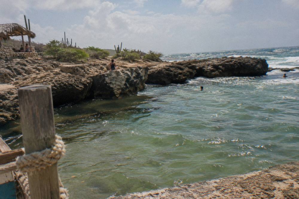 A rocky coastal natural pool with green rippled water, Atlantic surf, cactus-covered arid terrain and a thatched shade shelter