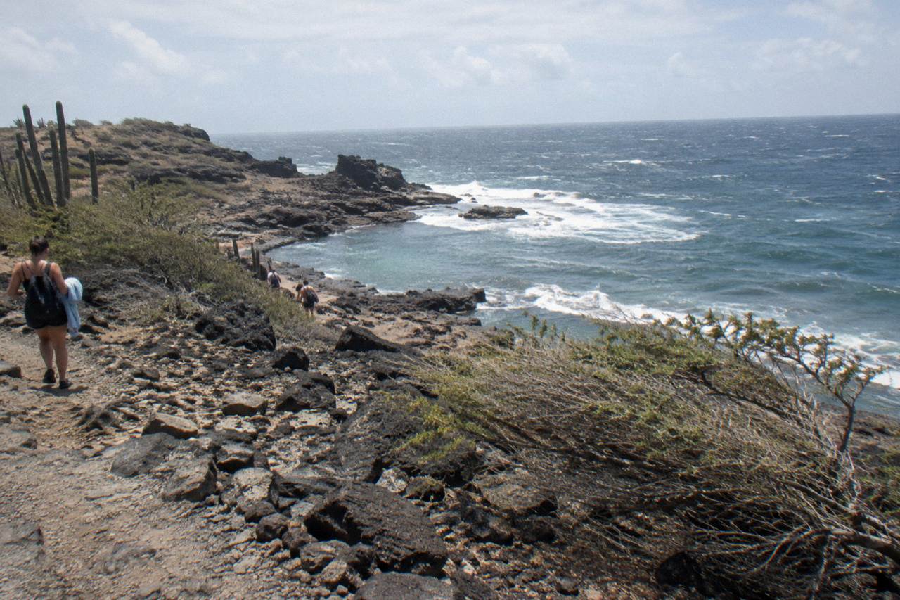 A rocky desert trail descending toward a natural pool set in dark volcanic rock on Aruba's north-east coast