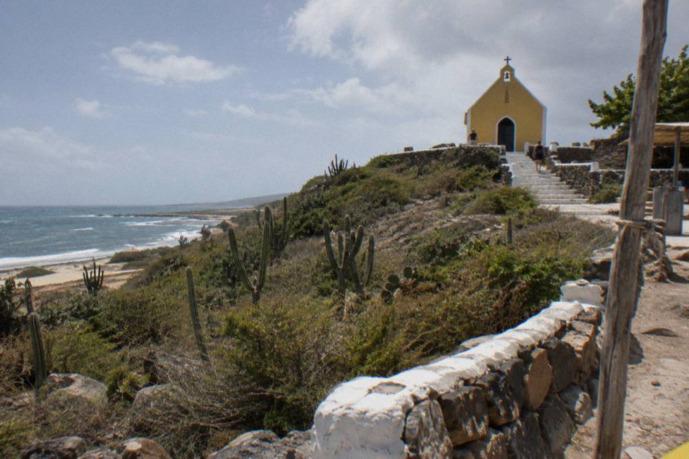 Yellow chapel on a scrubby coastal hill with cacti and ocean surf under a partly cloudy sky
