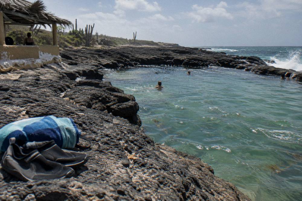 A pair of water shoes and a rolled towel set on volcanic rock beside a calm natural rock pool