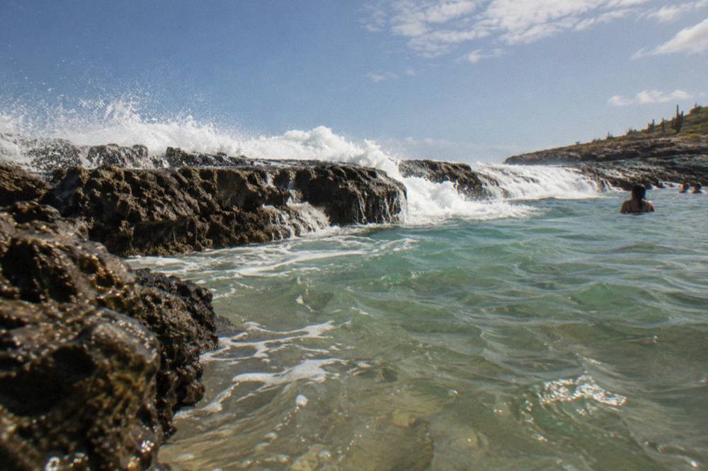 Heavy white waves breaking over a dark rock rim into a natural pool under a cloudy sky