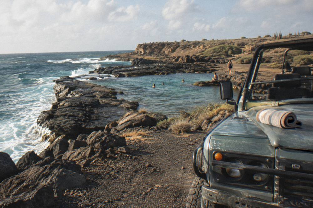 An open-air four-wheel-drive vehicle parked on a rocky track above a natural pool on the coast