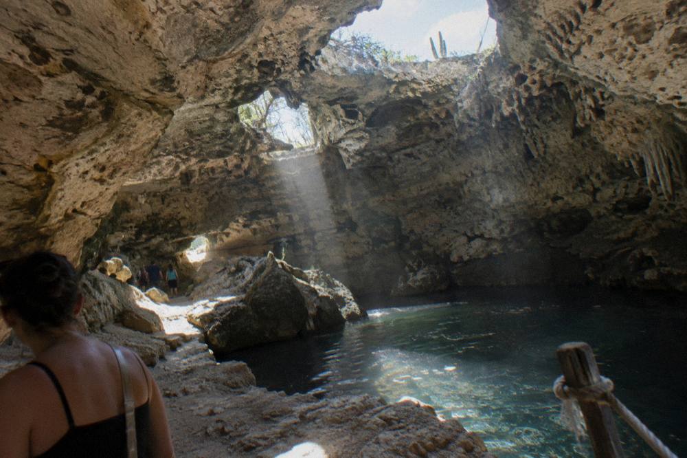 Limestone cave interior with sunbeams through ceiling openings, a dark pool, and rope railing beside visitors