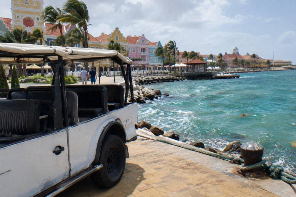 A white open-top jeep parked on a paved waterfront with turquoise water, rocks, palms and pastel Dutch-colonial buildings