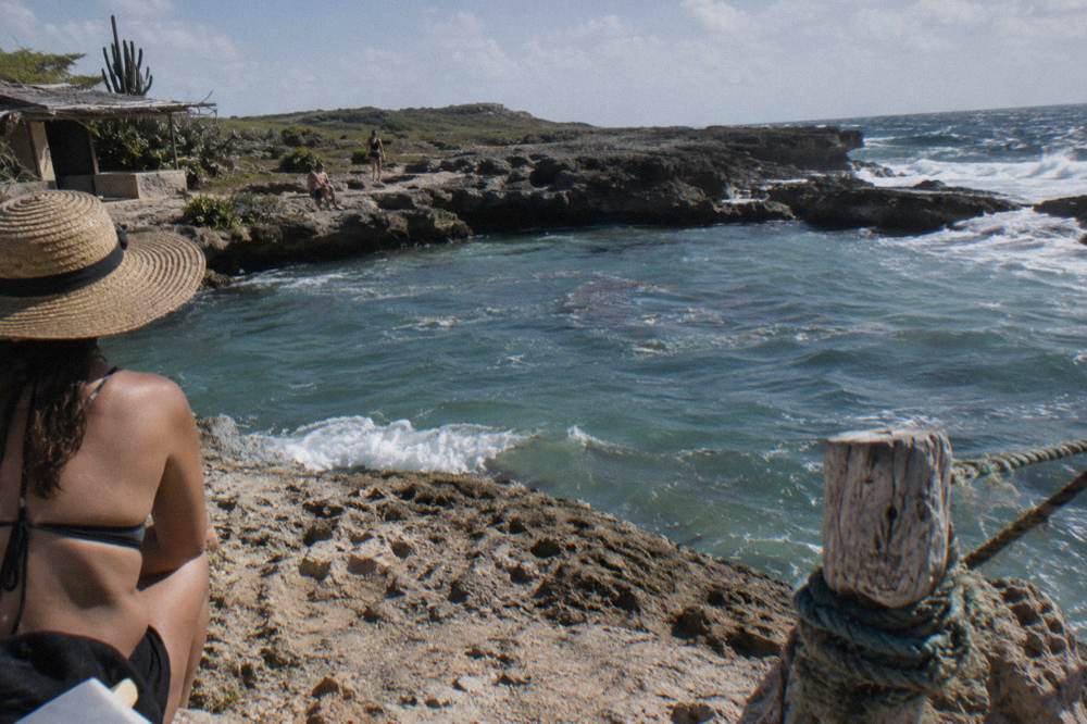 Person in a sun hat seated on the rim of a rocky natural pool with surf and cactus behind